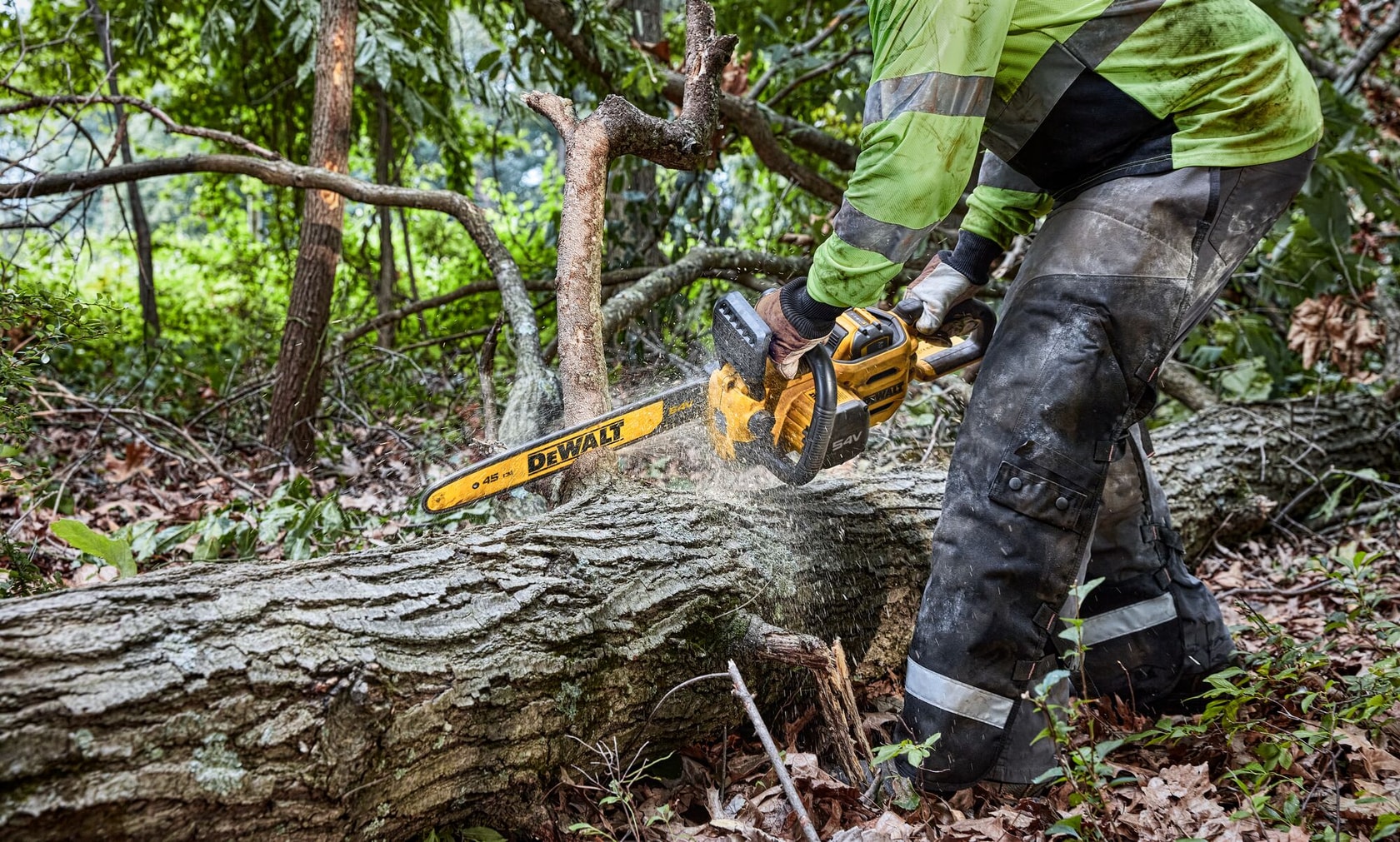 Uma pessoa com equipamento de proteção usa uma motosserra sem fio DEWALT para cortar uma árvore caída em uma área florestal.